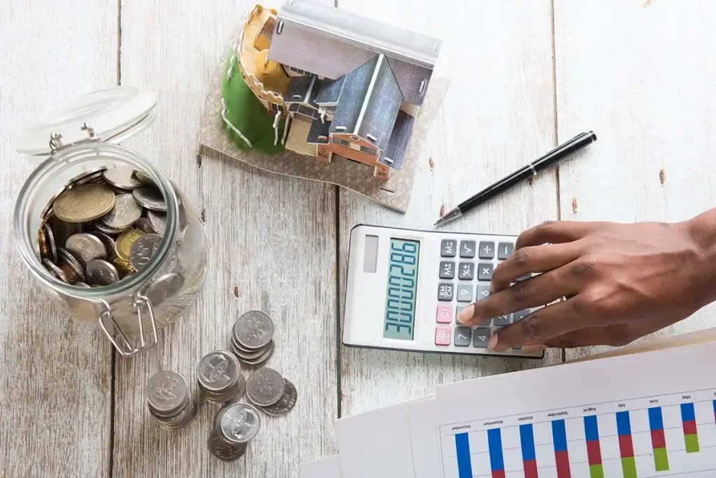 Model of a home, a jar of coins, printed graphs, and calculator on a table.