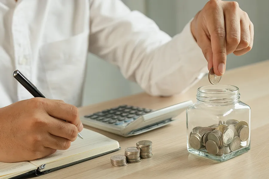 Man sitting with a calculator and a notebook, putting coins a jar.