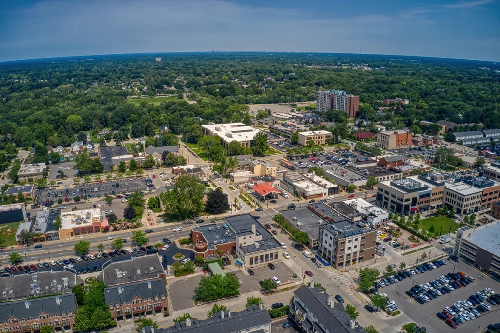 Aerial view of a suburban downtown area in Dearborn, Michigan, showing a mix of low-rise commercial buildings, office spaces, and residential neighborhoods. Streets are lined with parked cars, and the area is surrounded by lush green trees and dense foliage in the background under a clear blue sky.
