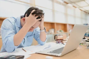 Man holding his head with both his hands in frustration. Papers scattered in front of him next to a laptop and a calculator.