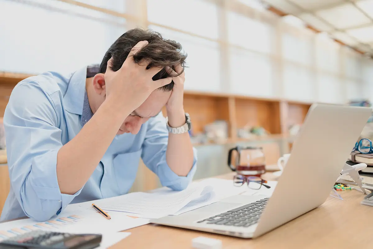 Man holding his head with both his hands in frustration. Papers scattered in front of him next to a laptop and a calculator.