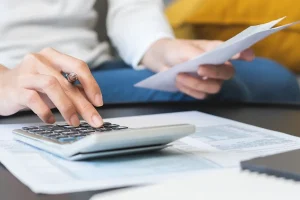 A person sitting at a table uses a calculator while holding a sheet of paper, reviewing financial documents spread out in front of them.