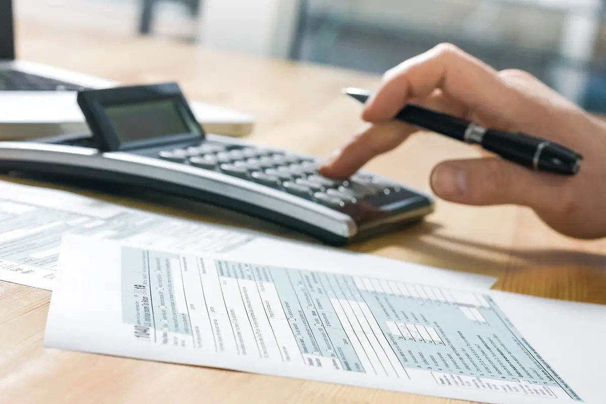 Hand typing into calculator while holding a pen on a desk with tax paper work on it.