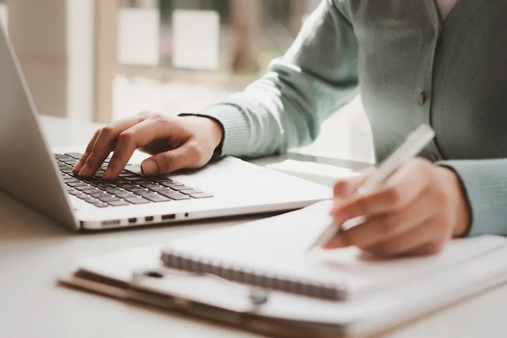 A persons hands, one typing on a laptop and one hand holding a pen on a notebook.