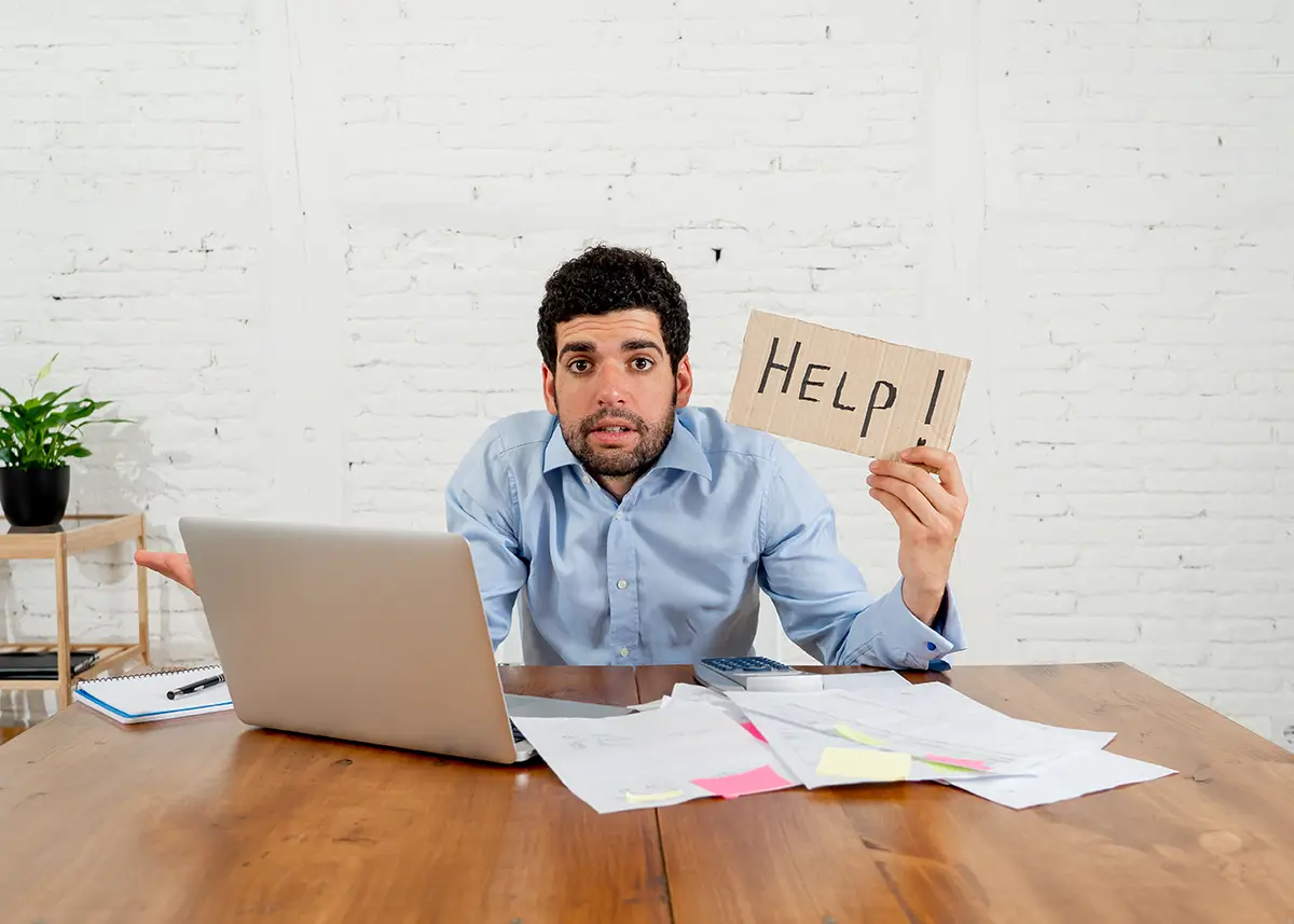 Overwhelmed young man trying to manage business taxes, holding up cardboard sign that says "Help"